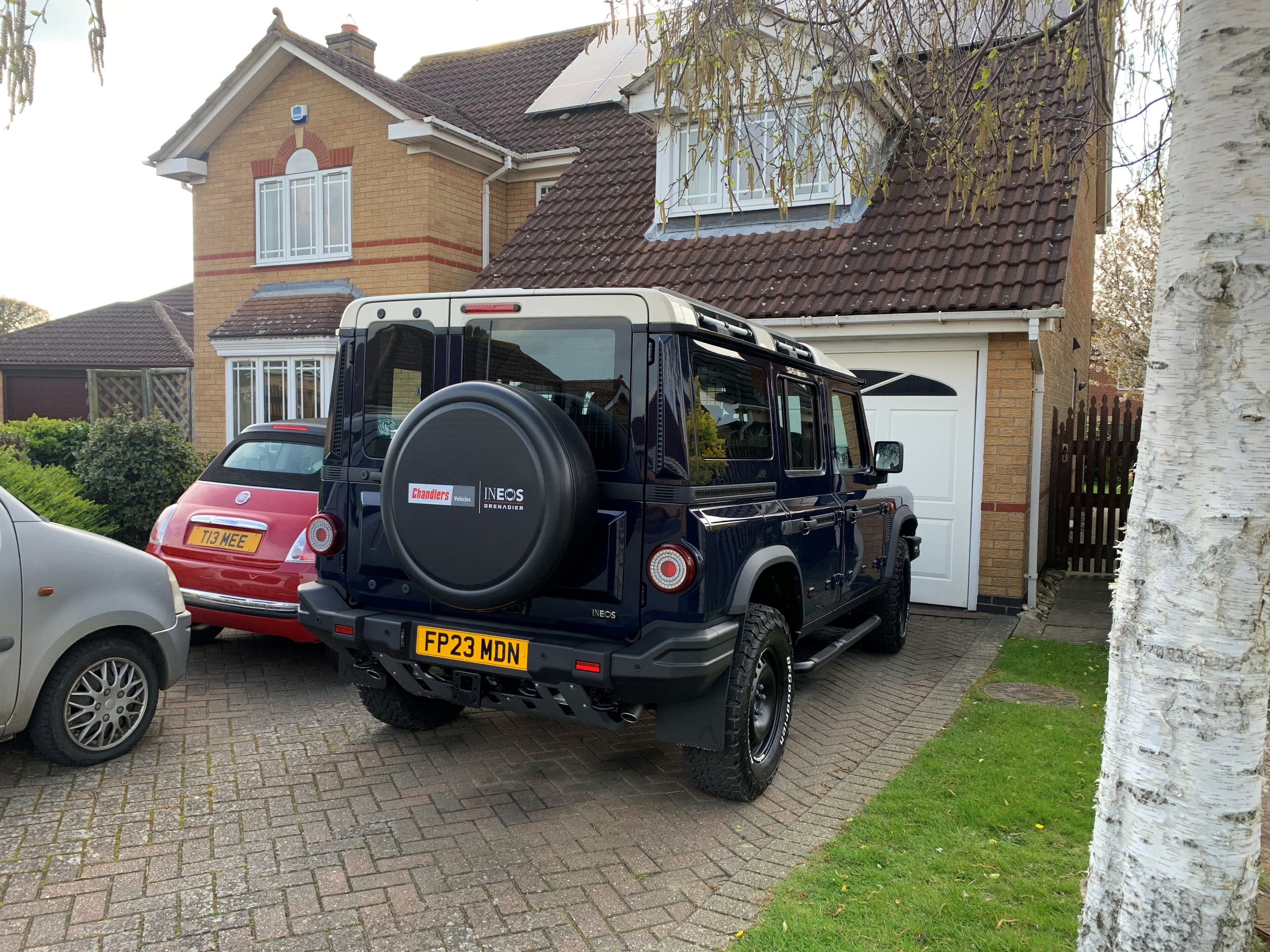 Ineos Grenadier customer cars arrive at dealers - Page 9 - INEOS - PistonHeads UK - The image shows a large, black SUV parked in front of a house. The vehicle is positioned at an angle, with the front left side visible. It has a distinctive red stripe on its door and features a spare tire mounted on the back. The house behind it appears to be residential, with a driveway and some trees indicating a suburban setting. There are no people or other moving objects in the scene. The focus is clearly on the SUV, suggesting it may be for sale or display.