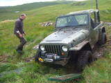 A man standing next to a large truck - Pistonheads - This image shows a rugged outdoor scene featuring an older, unused green off-road vehicle. A bald man dressed in a long-sleeved brown t-shirt is standing on a grassy hillside behind the vehicle. He appears to be examining the jeep closely, possibly checking for damage or maintenance issues. The vehicle is tilted to its left side, and there is some debris or material on the ground around the front tire, implying the jeep might have been driven through rough terrain. The terrain itself is lush and verdant, with shades of green and mild slopes. The image is a candid photograph, capturing a moment of utilitarian interest rather than a posed or artistic image.