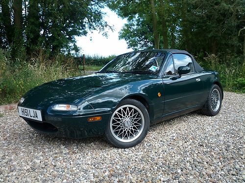 1991 mx5 limited edition- Rare i think pls help? - Page 1 - Mazda MX5/Eunos/Miata - PistonHeads - The image presents a kelly green sports car parked on a gravel lot. The vehicle has a sleek design, featuring two doors and a convertible top. A set of white-walled wheels with a distinctive silver rim is visible, and the license plate is partially visible, suggesting it might be registered in the UK. The car's setting appears serene, surrounded by a lush landscape with trees and greenery. There's no visible text or branding on the car, and it appears to be a sunny day, as indicated by the shadows being cast.