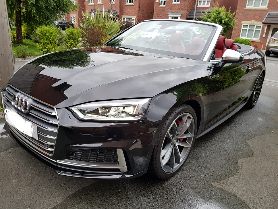 Looking to buy a S5 - Page 1 - Audi, VW, Seat & Skoda - PistonHeads UK - The image shows a sleek black Audi car parked on a wet surface, possibly after a rain. The vehicle is positioned at an angle that showcases its shiny exterior and the distinctive design elements of the Audi brand, such as the grille and headlights. There are no visible people or other objects in the immediate vicinity of the car, suggesting it may be parked on a street or in a parking lot. In the background, there is a partial view of what appears to be a residential area with houses and trees. The focus of the image is on the Audi, which is the central subject due to its prominence and detail.