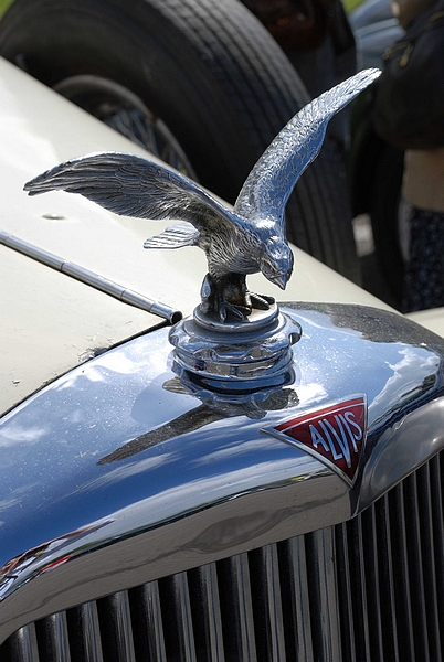 Goodwood Breakfast Club, Pre 1966 classic sunday - Page 1 - Goodwood Events - PistonHeads - The image showcases an old, classic car, prominently featuring the front grille which is illuminated under the sunlight, revealing its chrome finish. A notable detail is the emblem of the marque "Alvis" displayed on the grille, indicating the make of the vehicle. Perched on top of this ornate grille is a hood ornament, extinct by current standards, but characteristic of vintage automobiles. The hood ornament, depicting an eagle in flight, adds a touch of elegance and vintage charm to the car's aesthetics. The car appears to be the main subject of the photograph, with blurred elements in the background, suggesting the setting might be a classic car show or a similar event dedicated to the preservation and appreciation of such vehicles.