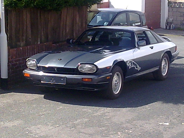 Newbie Pistonheads South West - The image shows a dark-colored sports car parked on a paved surface, seemingly on a street with residential houses visible in the background. The car's front end is prominently displayed, featuring two round headlights, a smaller black grille, and distinctive design elements typical of sports cars. The car is parked next to a white post or pillar that stands out against the vehicle's darker hue. There are no visible texts or distinctive brands in the image that are identifiable. The photograph has a casual, candid quality, likely taken without staging.