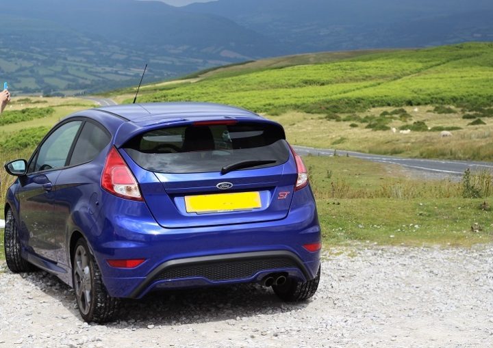 Fiesta St 180 - Page 5 - Readers' Cars - PistonHeads - The image depicts a vibrant blue Ford Fiesta parked in a gravel field with a picturesque backdrop featuring a hilly, grassy landscape and a blue sky dotted with clouds. The car is centered in the frame and appears to be owned by a woman, who is not fully visible; only the upper part of her torso is seen. She is holding an object in her hand, suggesting she might be interacting with the car or the surrounding environment. The car's license plate is clearly visible, indicating that the vehicle is likely from the UK or another region with similar vehicle registration formats. The overall setting appears to be a rural or semi-rural area, suggesting the location might be a countryside countryside.