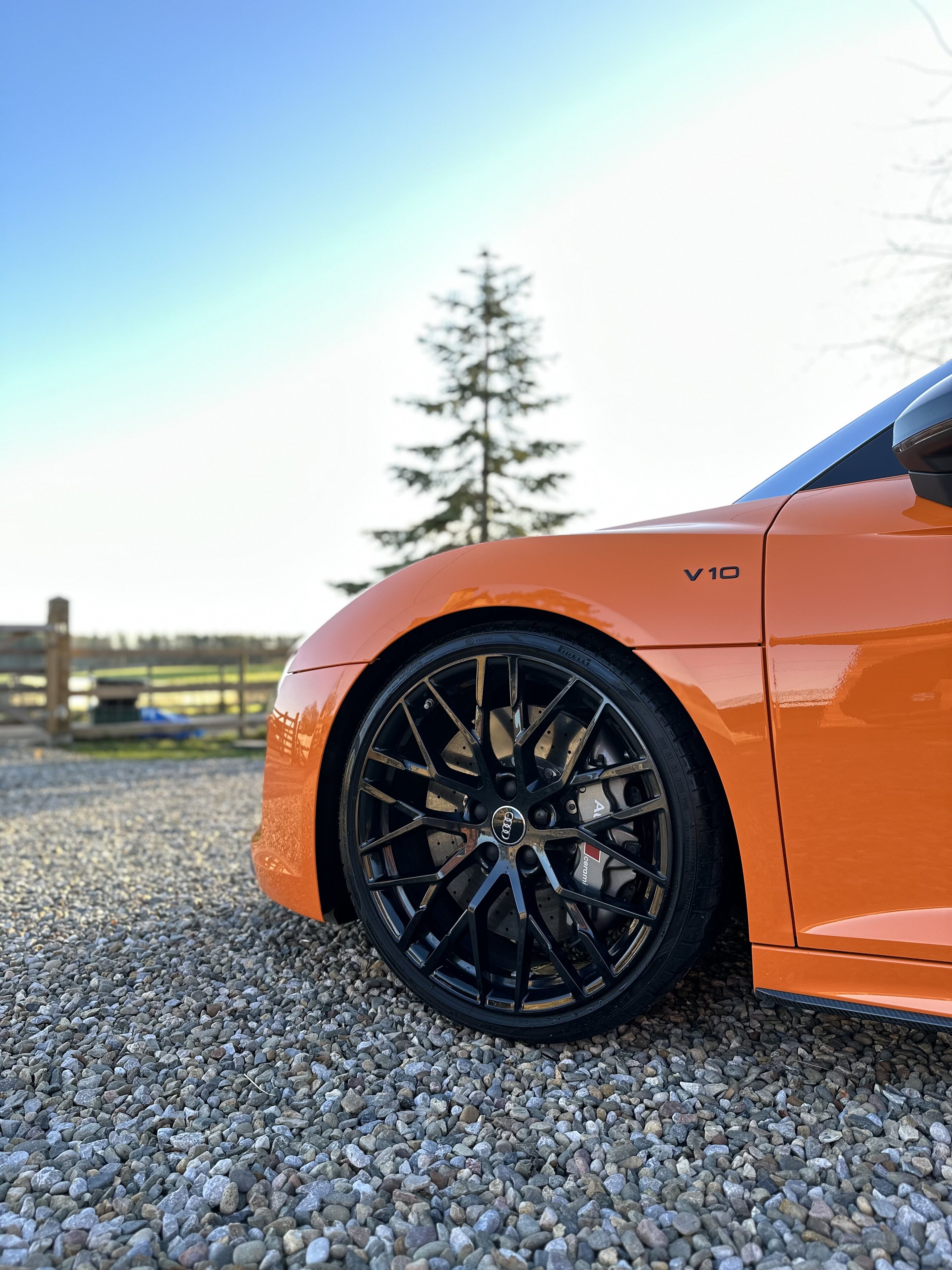 Orange Audi R8 V10 Plus Spyder  - Page 1 - Readers' Cars - PistonHeads UK - This image features a vibrant orange sports car parked on a gravel surface. The car has a sleek design with black rims and is positioned at an angle to the viewer. In the background, there is a clear blue sky and a hint of greenery, suggesting the car is outside in a rural or suburban setting. On the left side of the image, partially obscured by the vehicle, is a tree. The overall scene conveys a sense of speed and luxury associated with high-performance sports cars.