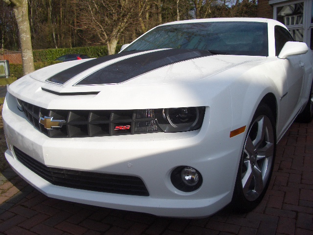 2010 Camaro - Page 1 - Yank Motors - PistonHeads - The image features a white Chevrolet Camaro ZL1, a high-performance sports car, parked on a brick driveway. The car's front facia is clearly visible, showcasing its sleek design, large air intakes, and a prominent grille logo. The Chevrolet bowtie is prominently displayed on the car's grille. The setting is outdoors with trees and a partially visible house in the backdrop. The overall atmosphere of the picture suggests a residential area with the car parked near a residence. There are no people present in the scene.