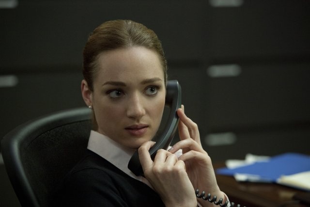 In this image, I see the acclaimed actress Emilia Clarke, known for her role as Daenerys Targaryen in Game of Thrones. She is shown in a moment of deep thought or concern, her right hand still holding the phone receiver to her ear while her left hand has fallen to her chin in contemplation. Emilia is seated at a desk, suggesting a professional or office setting. The backdrop is softly focused, drawing the viewer's attention mainly to Emilia, dressed in a no-nut gray jacket and a high collar blouse, which adds a touch of formality to her appearance.