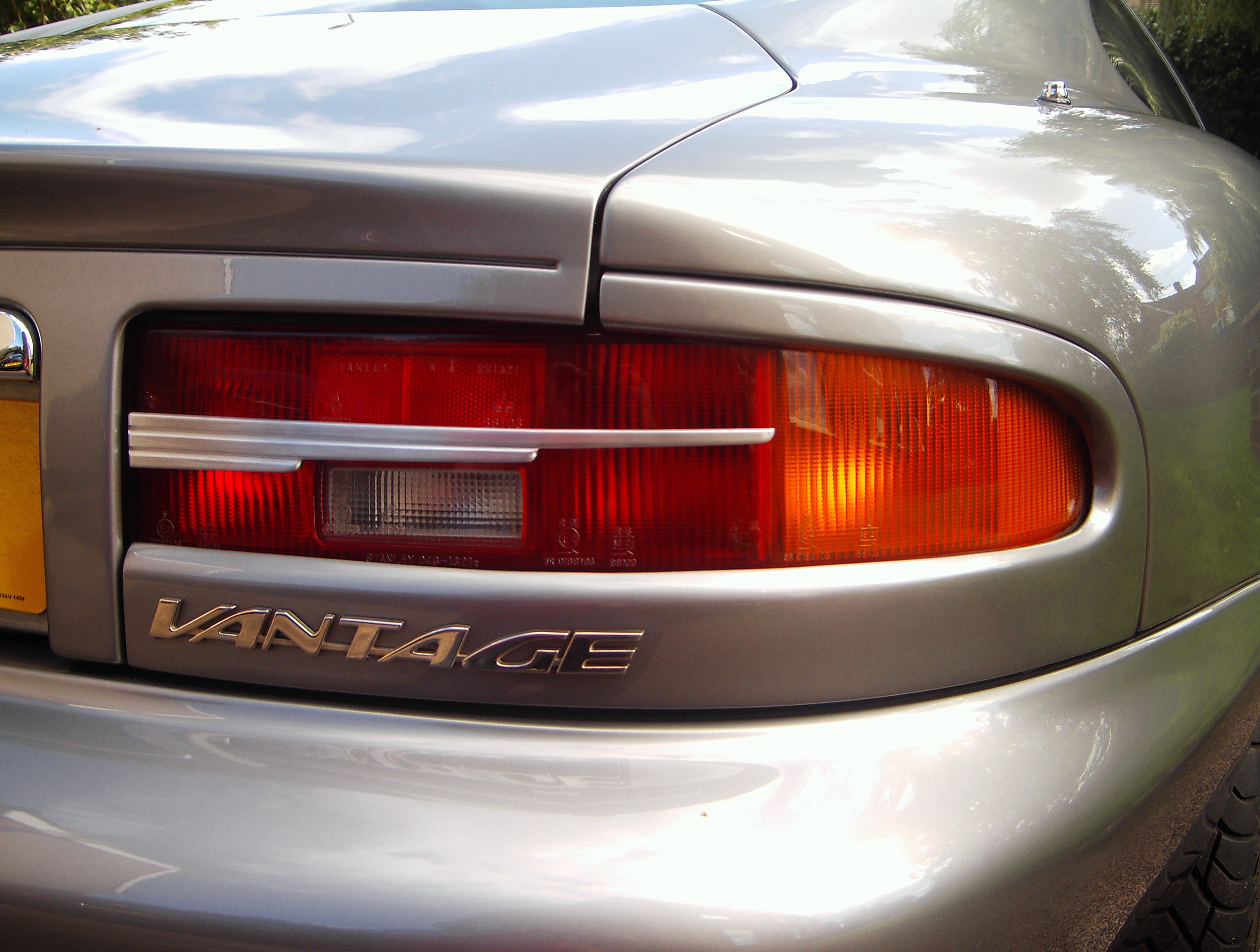 DB7 Rear Lights - modifications - Page 2 - Aston Martin - PistonHeads - This is a close-up photograph of the rear tail light of a vehicle. The tail light appears red, tinted by the vehicle's own diffuser. The brand of the vehicle, "VANTAGE", is prominently displayed, indicating the make. This image is taken during the day with ample natural light, which casts highlights on the vehicle's shiny surface, emphasizing its smooth and clean appearance. The photo captures the poised stance of the vehicle, its red tail light illuminated, ready for action or simply parked and waiting.