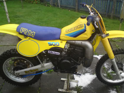 Show us your 2 stroke prawn - Page 7 - Biker Banter - PistonHeads - The image shows a Suzuki dirt bike parked on what appears to be a wet surface, possibly a driveway. The bike's frame is a vibrant yellow, and it features the letters "R&M" in a bold blue color on a blue seat. The bike's number "500" is also visible. The front wheel is mightily dirt accumulated, suggesting the bike has recently been used. The setting is a quiet, suburban yard with a green lawn and a wooden fence in the background.