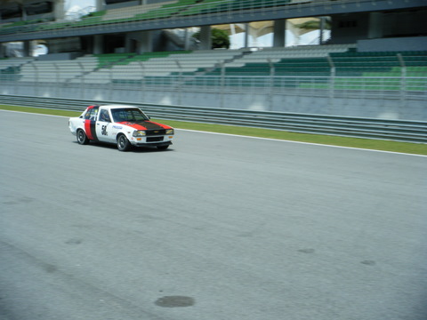 Pistonheads Afos Sepang - This image shows a scene at a race track. The focal point is a car, predominantly painted in a red, white, and black color scheme, likely with racing sponsors' branding, speeding down the track. The car is positioned in the lower third of the image, moving towards the right. The photograph captures motion, creating a sense of speed. The setting includes a motor racing course with a pavement, a large, empty spectator stand with benches on the left side of the image, which is built below the foreground. The infrastructure and the presence of the car suggest this could be a professional racing event.