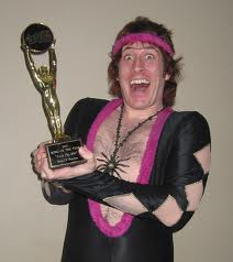 The Football Forum/MOTD/SS/Transfer/General Rant/OT thread - Page 84 - Football - PistonHeads - The image shows a man dressed in a theatrical costume, which includes a black outfit with highlights of pink and gold sequins. He is wearing an elaborate headband and has wild hair. The man is holding a trophy in his right hand, which he is displaying with pride. The trophy is a statue of a person reaching up, almost as if to touch the sky. The background is a simple, plain wall. The man's expression is joyful, and he appears to be momentarily overwhelmed with happiness. The overall impression is that of a performance or theatrical presentation.