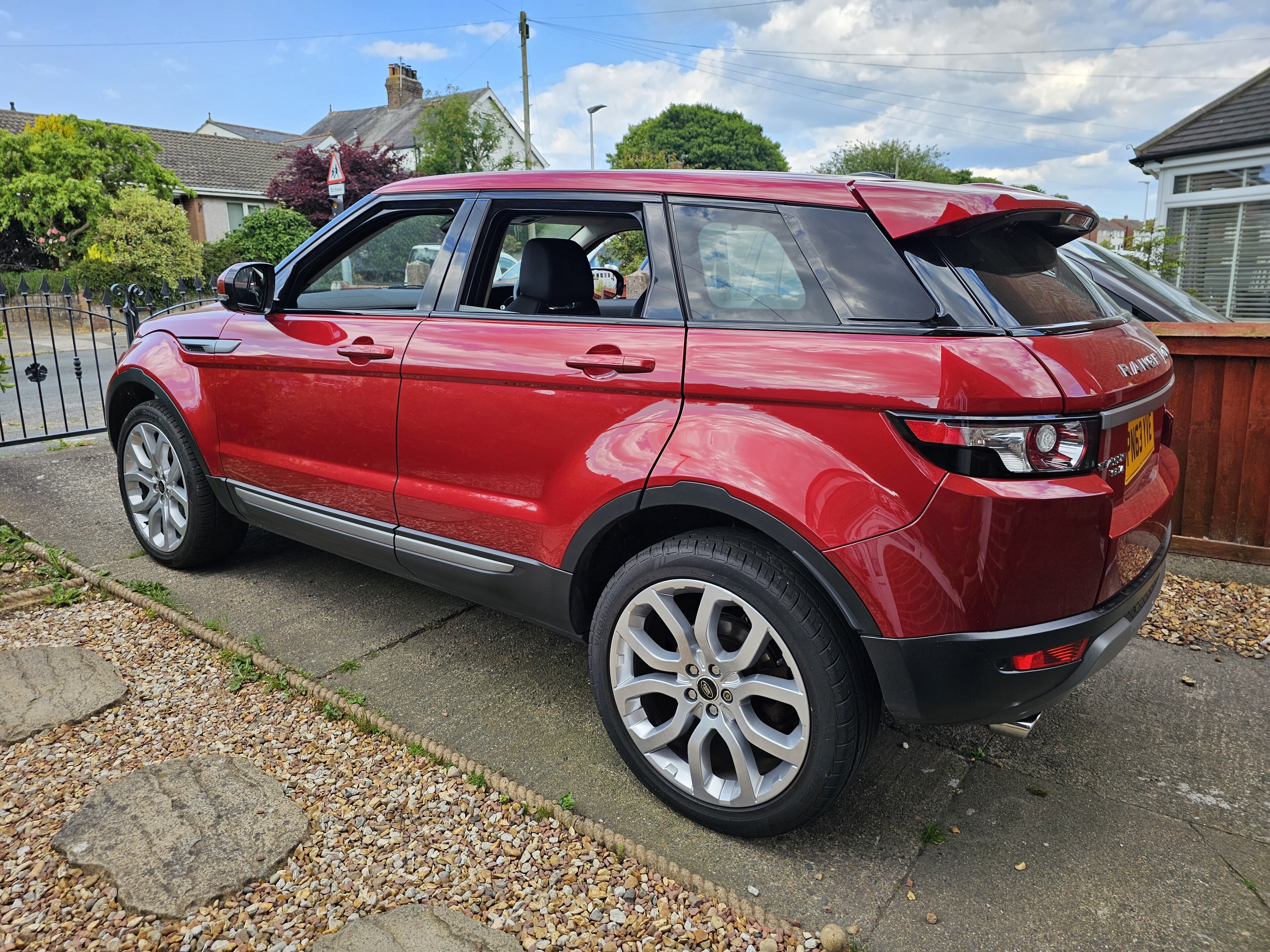 SUV Advice needed - Page 1 - Car Buying - PistonHeads UK - The image shows a vibrant red Range Rover parked on the side of a road. It's a large SUV, featuring black roof rails and silver wheels. The vehicle is positioned next to a fence and a hedge, suggesting it might be outside someone's home. There are no visible people in the picture. The photo appears to be taken during the day under clear skies.