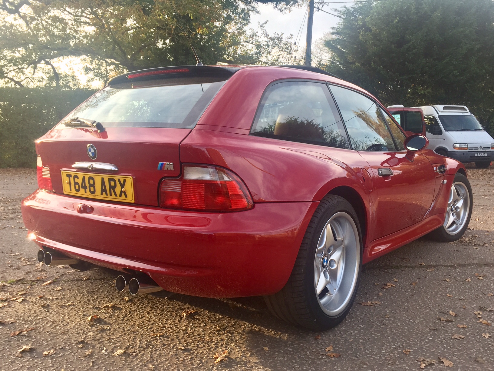 RE: BMW Z3 M Coupe: Spotted - Page 3 - General Gassing - PistonHeads - The image shows a vibrant red sports car parked on a paved area next to a well-pruned green bush. The backdrop of the scene reveals a field, a few scattered trees, and another vehicle parked at a distance, contributing to an open and serene outdoor setting. The car itself is equipped with a prominent rear spoiler, large circular rims, and a noticeable license plate. The overall composition creates an atmosphere of tranquility, with the red sports car as the center of attention.