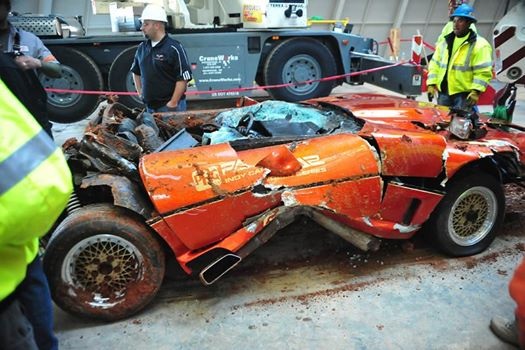 1,000,000th. Corvette returns from chasm - Page 1 - Corvettes - PistonHeads - The image shows a scene with a severely damaged red sports car situated in the foreground. It appears that the car has been involved in a high-speed accident, exhibiting crumpled and bent metal, shattered windshield, and misaligned body parts. The car is sitting on a track or road, and there are several people looking at it, indicating it might be the aftermath of a race or event. In the background, there's a truck parked, and an individual in high visibility clothing is standing nearby. The overall setting seems to be a industrial or industrial-like environment, possibly a repair or inspection facility. The colors in the image are quite vivid, with the red of the car contrasting against the grays of the other objects and the blue of the ground or floor.