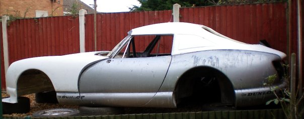 Cerbera Style Hard Top for Chimi´s - Page 4 - Chimaera - PistonHeads - The image depicts an old-fashioned silver car parked in a yard behind a red fence. The car appears to be an older model, likely from the 1950s or 1960s, given its design. It's leaning slightly to the left, possibly from age or lack of support. The setting suggests a suburban or rural area, with the fence and bare ground indicating an outdoor, possibly unkempt, location.