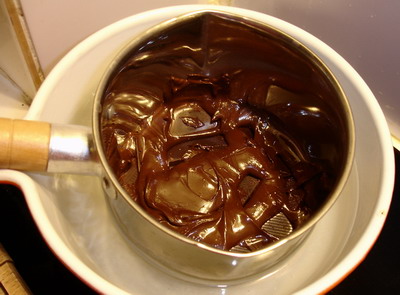 The image captures a close-up view of a process where melted chocolate is being poured into a pan. The chocolate has a rich, dark color and is in the midst of being poured from a spouted container, likely a bowl or pot. The melted chocolate appears silky and smooth, and some of it is visible dripping into the same container it's being poured from, indicating that some has already been poured. The surface holding the container appears flat, possibly a table, which contrasts with the sides that are out of focus. The focus of the image is on the chocolate, its smooth texture and rich color, as it is the main subject of the photograph. There are no visible texts or distinctive markings that provide further context.