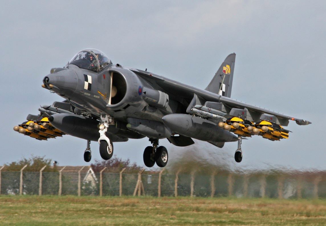 A fighter jet flying through a blue sky - Pistonheads - The image captures a dramatic scene in the sky, featuring a military jet flying low over a grassy field. The jet is predominantly dark gray, with distinctive markings on its grand wings. The aircraft's nose is pointed downwards, suggesting a high-speed maneuver. Below the jet, there are external structures which appear to be weaponry or payloads, ready for deployment. The backdrop of the image is a hazy sky, adding to the intensity of the scene. This appears to be a test flight or demonstrative exercise, as evidenced by the strategic position of the military jet and the precision-like movement of the aircraft.