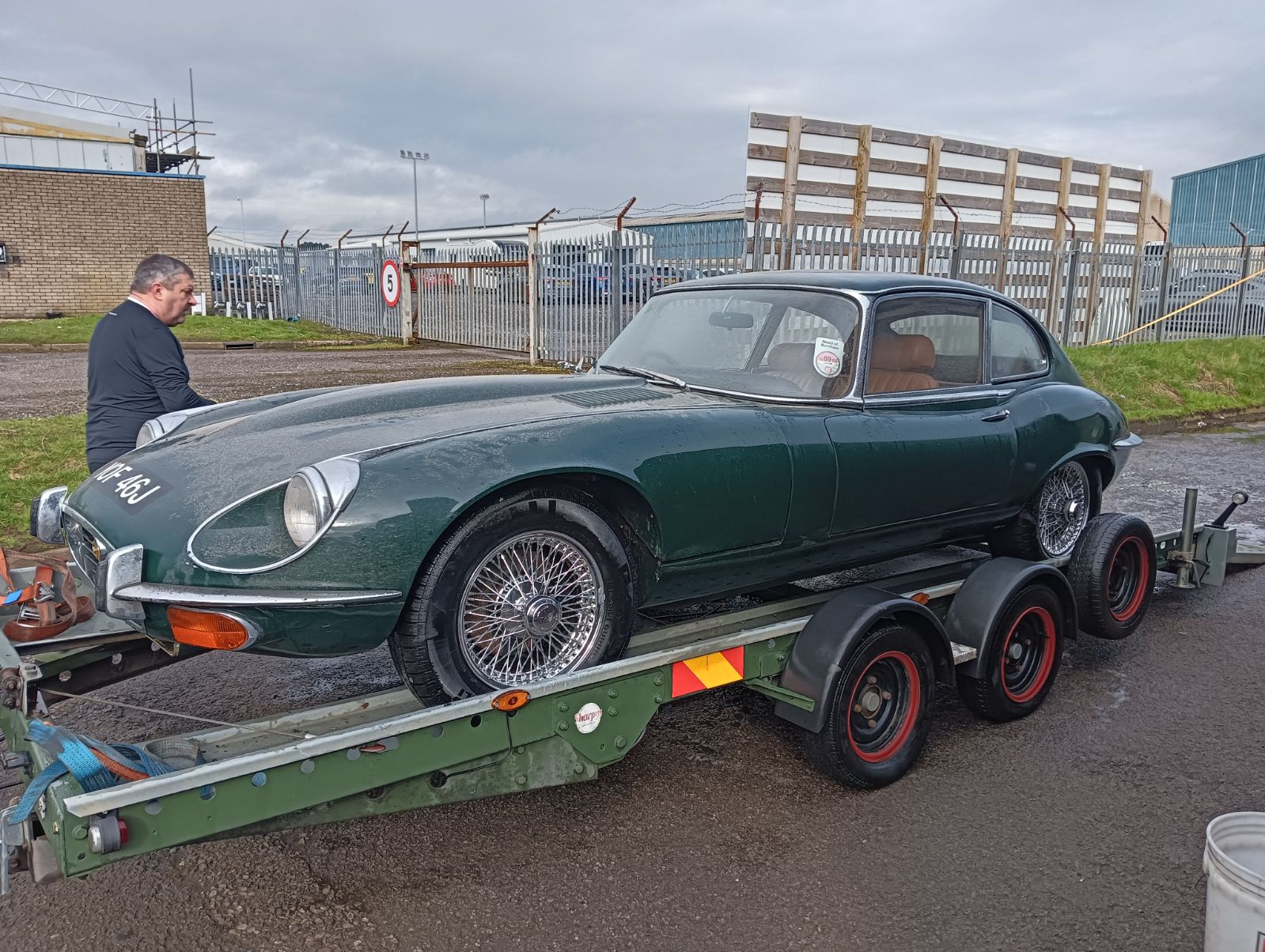 Pistonheads - The image shows a vintage car being towed on a trailer. The car is green and appears to be an older model, possibly an Aston Martin. It's attached to a flatbed trailer, which is hauling it. There are two individuals present in the scene; one is standing next to the trailer while the other is positioned further away. They seem to be engaged in some activity related to the car or its transport. The setting suggests an outdoor environment with overcast skies, and there's a building visible in the background.