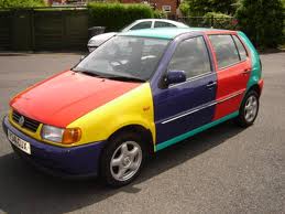 Pistonheads Interior Kestrel - The image shows a car parked on an asphalt surface. The car is decorated with several sections of different colors. It had a red base color on the bottom, a colorful central section with yellow, black, and light blue, and a white top. It has a visible licence plate. In the background, there is a building, which might be residential, and a suburban road. The car has a distinctive design, possibly suggesting it is a custom or modified vehicle.