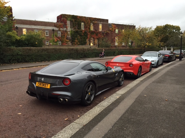 Supercars spotted, some rarities (vol 6) - Page 321 - General Gassing - PistonHeads - The image shows a line of cars parked alongside a curb on a city street. A striking red sports car is parked in the foreground, adjacent to a metallic grey exotic sports car. These two cars are positioned next to each other along the curb. In the background, there are more cars and a building with a brick exterior. The setting appears to be in the evening under an overcast sky, as suggested by the muted lighting. There is a person and a bicycle nearby, adding a touch of everyday life to the scene. The cars' sleek design and shiny exteriors hint at their high-performance nature.