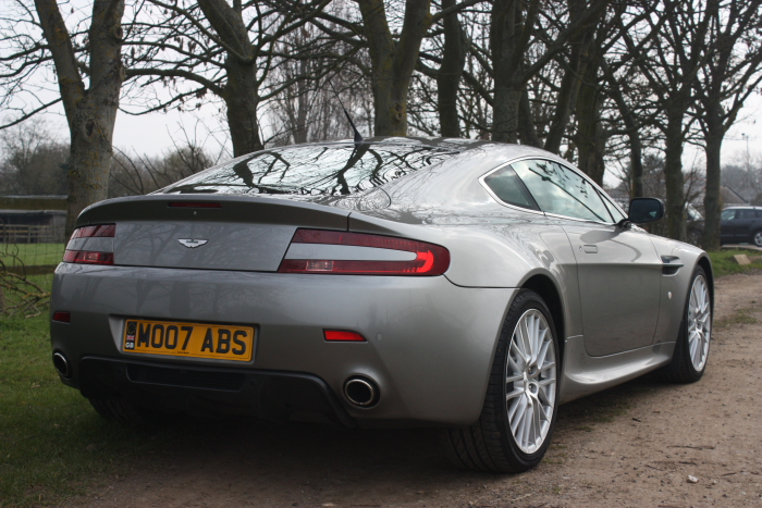 Pistonheads Difference Spot - This image features a sleek, gray sports car parked on a gravel path. The car, bearing the license plate "M007 AB5," is an Aston Martin DB7, characterized by its round emblem and racy design. The setting is picturesque, with the car in front of a line of tall, leafless trees, suggesting a late autumn or winter season.