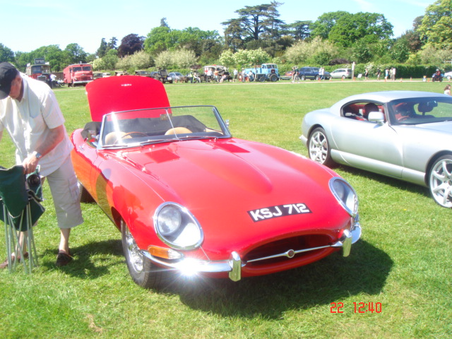 Pistonheads - The image depicts a vibrant scene at an auto show. The red KJS 712 sports car is parked in a grassy field adjacent to two other race cars. The sports car's shiny red finish contrasts beautifully with its silver wheels. On the sidelines, a man is seen in a white T-shirt, attentively observing the cars with a relaxed posture, his hands casually resting on a well-equipped green motorcycle. The atmosphere is charged with excitement and fascination for the beautiful machines on display.