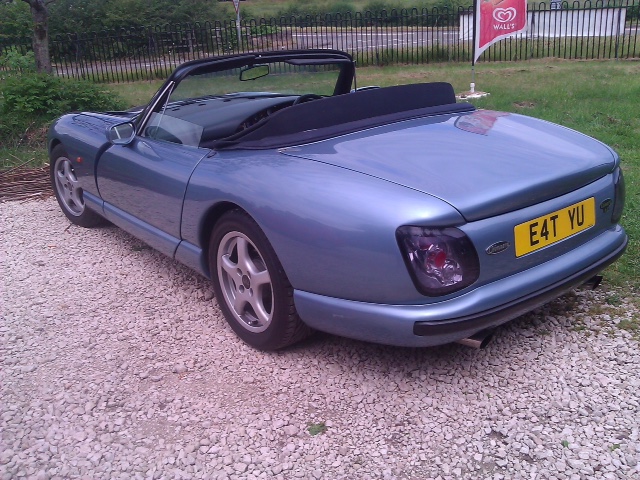 TVR Number Plates Love 'em or loath 'em there's plenty - Page 1 - General TVR Stuff & Gossip - PistonHeads - The image shows a blue convertible sports car parked on a gravel area. It's a sunny day, suggesting the photograph was taken during the summer. The car features a black soft roof that is closed. In the background, there's a lush green lawn and a wooden fence. There's also a small red flag on the car, adding a pop of color to the scene.