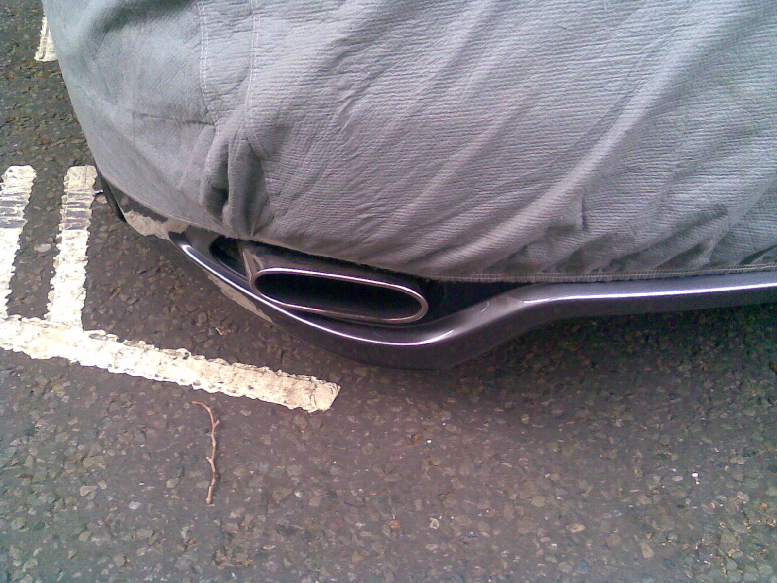 Ultimate Car Spotter Thread - Page 1 - General Gassing - PistonHeads - The image shows the back of a vehicle parked on a street. A spare tire is securely fitted under the car and is covered with a gray canvas cover. A white stripe, possibly a parking line indicator, runs along the road beside the tire. The car appears to be a modern model, and the spare tire area is relatively unobtrusive. The scene suggests a residential area as it's in shades of gray, likely due to the weather or lighting conditions. There are no people visible in the image.