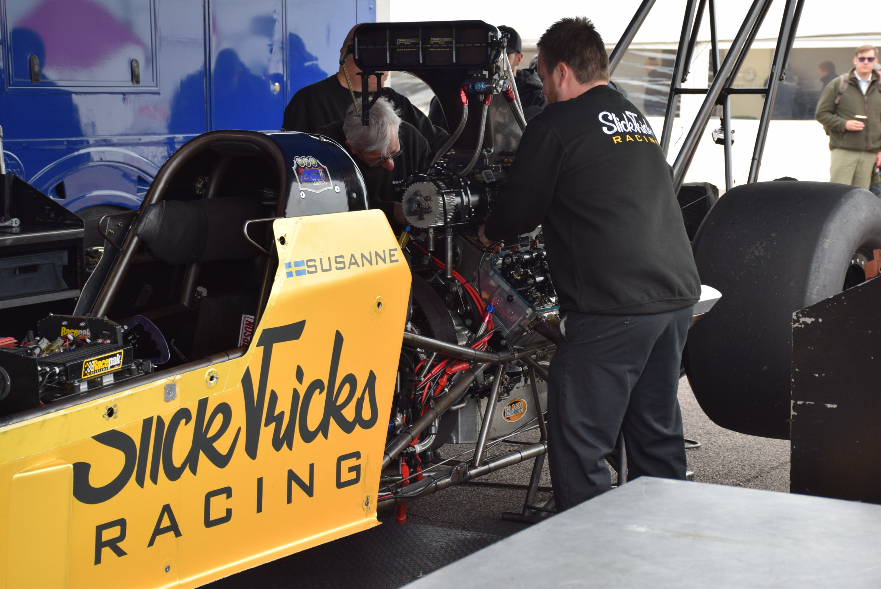 Pistonheads - The image shows a man working on a race car. He is standing next to it, with his hands on the engine cover, which displays the name "SICKSTICKS RACING." The car has a yellow and black color scheme, and there are various tools and equipment scattered around the area, suggesting an ongoing setup or maintenance process. In the background, there's another person partially visible, and a truck is parked nearby.