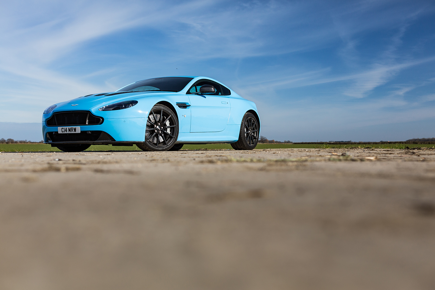 So what have you done with your Aston today? - Page 299 - Aston Martin - PistonHeads - This image showcases a striking blue Aston Martin DB11 sports car standing on a dirt surface with high clouds visible above. The car's sleek design combined with its vibrant color make it the focal point of the scene. In the foreground, there is a small mound of sand, and the adjacent background reveals an open field under a vast sky. The landscape and the car's color contrast sharply, adding depth and visual interest to the image.