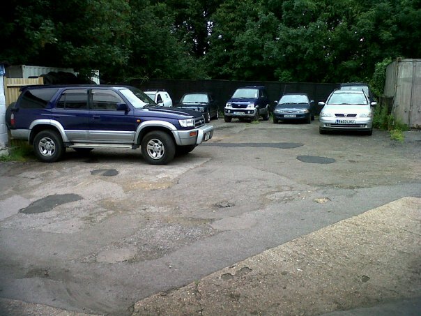 The BAD PARKING thread - Page 394 - General Gassing - PistonHeads - In the image, a group of cars and trucks are parked in what appears to be a lot, with trees quite close to the lot. The most prominent vehicle in the image is a blue SUV. The lot has a mix of different models and types of vehicles, including cars and trucks. The presence of a fence in the background suggests that the lot might be positioned in an area that requires some boundary. The trees surrounding the lot introduce a touch of nature in the otherwise hard, asphalt lot.