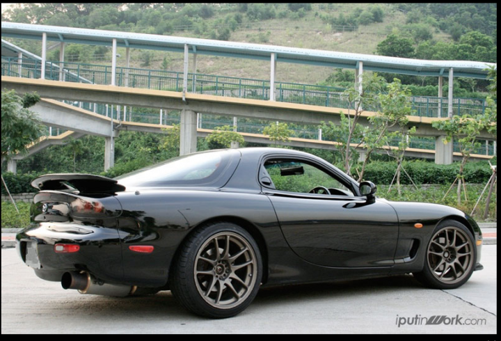 92' RX-7 Rebuild - Page 1 - Readers' Cars - PistonHeads - The image captures a black sports car, a convertible with a rear spoiler, parked on the street next to a nature trail. The car is positioned in the foreground, its sleek design accentuated by the roll bar that rises just behind the seats. The setting is serene, with the trail leading into a lush forest filled with verdant trees and bushes. An overhead bridge or arc spans the scene, suggesting a connected network of roads. There is a sense of solitude, with no immediate buildings or other objects in sight, reinforcing the feeling of escape from the city. The car's presence implies the anticipation of an exhilarating drive through the natural beauty that lies ahead.