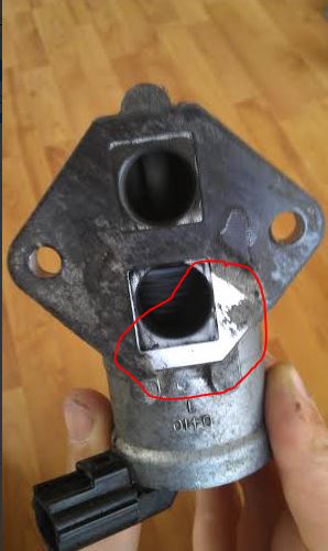 Yet again problems... engine stutters and nearly stalls - Page 1 - Mazda MX5/Eunos/Miata - PistonHeads - The image shows a close-up view of a mechanical component, likely a hose fitting. The component appears to be made of metal with visible wear marks and rust. There's a red circle drawn around a hole within this fitting. Below the fitting, there's a black component with text on it, although the text is not visible in this image. The component is placed on a wooden surface. The image focuses on the interplay between the red circle and the hole within the metal fitting, drawing attention to a finer detail.