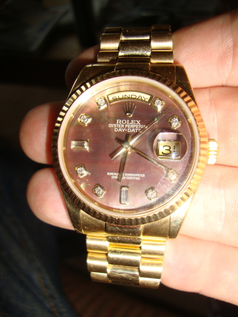 Pistonheads - The image shows a close-up view of a person's hand holding a luxurious Rolex 'Sunday' day-date wristwatch. The watch has a shiny gold band and an intricately textured dial with Roman numerals. The date on the watch is clearly visible as '31'. There is a crown on the right side of the watch face, indicating the area used to adjust time settings. The overall image presents a sense of elegance and luxury setting.