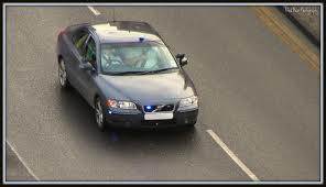 Suspicious white Skoda - M5 Cheltenham area - Page 1 - South West - PistonHeads - This is a color photograph showcasing a gray-colored vehicle driving away on a blacktop road. The car features a noticeable blue light on its front, which may indicate that it is either an emergency vehicle or a taxi. The road has white lane dividers, and the right side of the vehicle has a visible license plate. The vehicle is seen from a distance, as evidenced by the slightly blurred and elongated appearance, adding a sense of motion to the scene. The backdrop is relatively nondescript, focused primarily on the road and the vehicle.