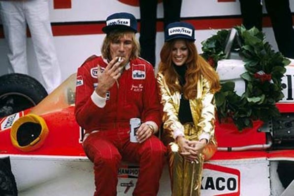 A group of people that are sitting on a bench - Pistonheads - The image features a male and female rider, presumably race car drivers, sitting together on a red race car. They appear to be from a past era, judging by their attire and the style of the car. The driver on the left is in a red racing suit with the number 7 on it and is smoking a cigarette while smiling at the camera. His outfit includes the name "Hamilton" visible. The driver on the right is in a shiny gold racing suit with "KACO" visible, also smiling. Both are wearing matching red, white, and blue driver's caps, and there are floral decorations on the car. They hold a bushel of wheat, and a can of beer is placed nearby.