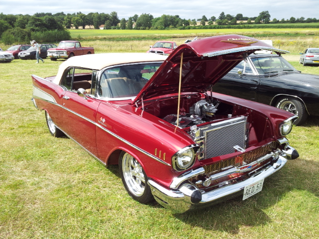 NSRA Hot Rod Supernats - Page 1 - Yank Motors - PistonHeads - The image features a vintage red car with the hood wide open, revealing its engine compartment. The car is parked on grass in an outdoor setting, where it stands out prominently against the natural backdrop. There are other cars in the background, but the focus is clearly on the vintage red car. The car's design suggests it's an older model, possibly a Chevrolet Nomad given its characteristic roofline and deck lid.