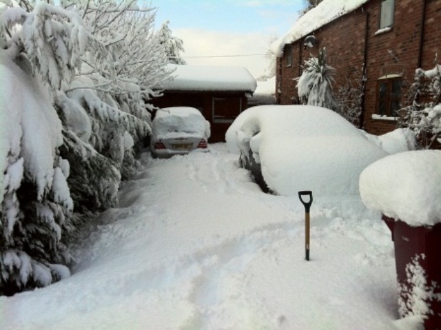 3 points & fine for snow on roof  :) - Page 2 - General Gassing - PistonHeads - The image depicts a heavy snowfall around a residential area. Cars are buried under deep snow, making it difficult to navigate the driveway. On the left side of the driveway, a snow shovel is visible, suggesting attempts to clear the snow. The driveway slopes upwards towards a brick house on the right side of the image, hinting at the effort required to reach such an elevated destination in the snow. The scene conveys the logistical challenges posed by snowfall in certain regions.