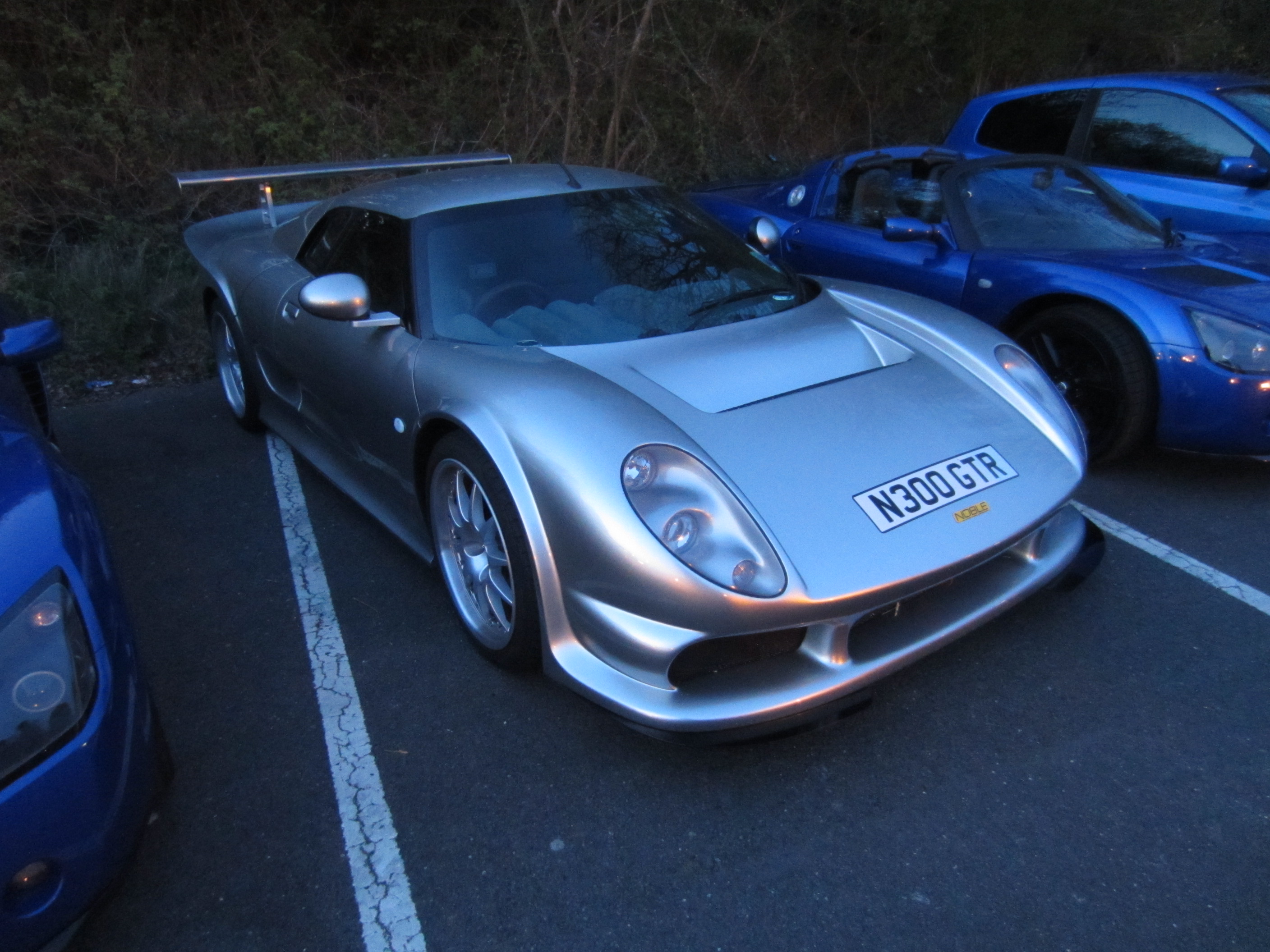 A car with a surfboard strapped to the back - Pistonheads - The image shows a parking lot with a focus on two sports cars. The first is a silver sports car, parked prominently in the middle of the space with crisp lines and shiny finish suggesting it's well-maintained. The second car is parked in the background on the right side, partially hidden by the silver car. It appears to be a similar model, also a sports car, painted in a blue hue. There are visible license plates on both cars. The parking lot itself is paved and painted with white lines, indicating it's a public or commercial parking area. The lighting in the image suggests it's either early morning or late evening, emphasizing the sleek design and glossy finish of the cars.