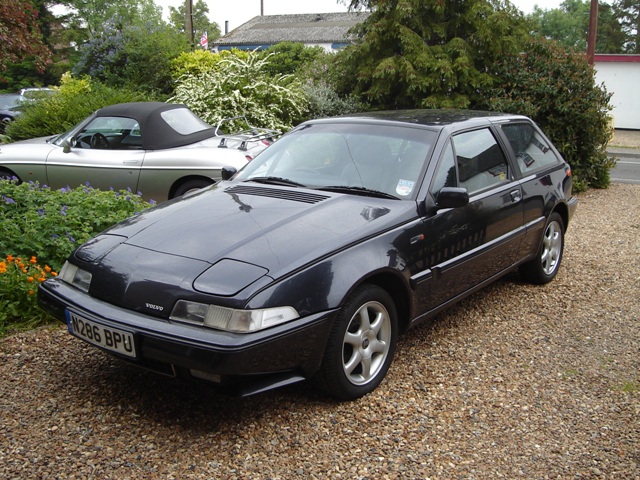 Pistonheads - The image shows a dark-colored hatchback parked on a gravel driveway. The car has a distinctive front end design, featuring a raised hood and a sleek body that reflects its quality craftsmanship. The gravel driveway is bordered by an array of plants and trees, adding a touch of greenery to the scene. In the background, another car can be partially seen, suggesting the presence of a neighboring house or driveway not visible in the frame. The overall scene presents a tranquil suburban setting, showcasing the car prominently as the main subject.