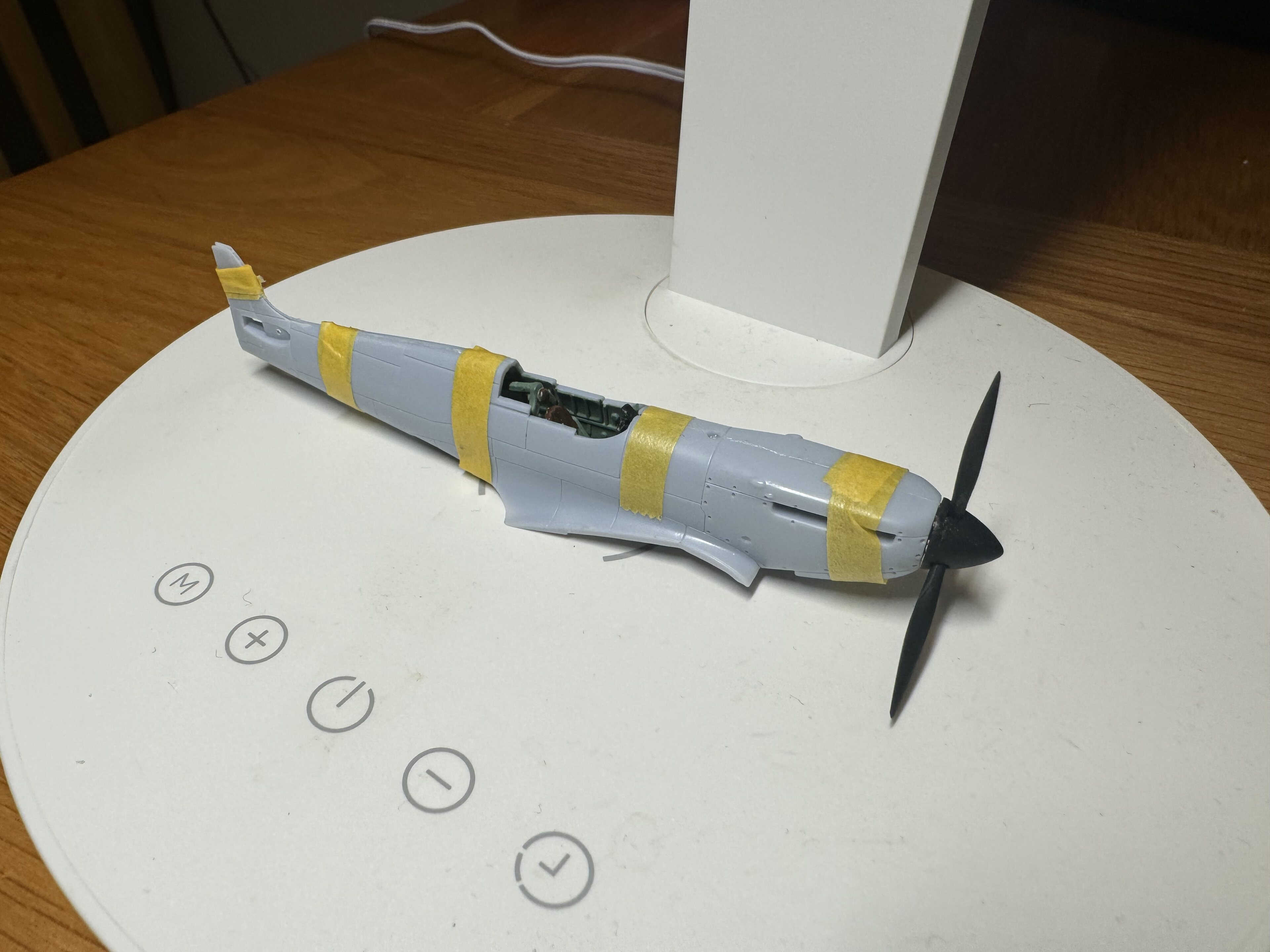 Pistonheads - This is a photograph of an intricately crafted model airplane displayed on a round, white stand. The plane is predominantly white with yellow markings and has a propeller on the front. It appears to be a representation of a fighter jet given its small size and stylized design. There are four buttons arranged in a circle beneath the stand, which might suggest interaction or control elements. In the background, there's a glimpse of a wooden table and a partially visible object that resembles a toy car. The overall scene suggests a hobbyist setup for displaying and possibly controlling these models.