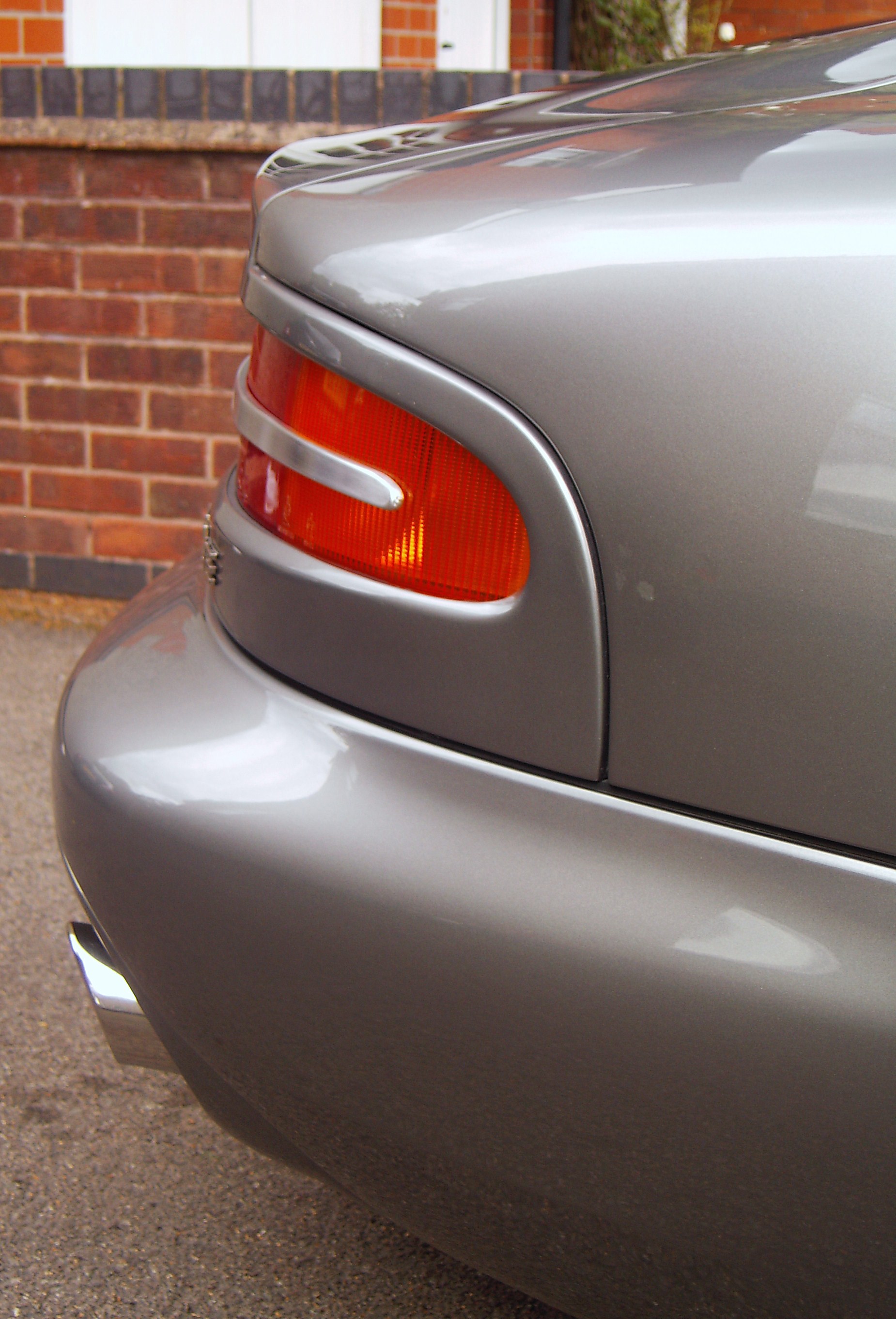 Lights Pistonheads Modifications - A silver, late-model car is the main subject of the image. The car is parked on the side of a road or driveway, adjacent to a red and black brick wall. A single red tail light is visible on the back of the car. It is a clear day with ample natural light illuminating the scene. There is one person somewhat visible in the background to the left side of the image.