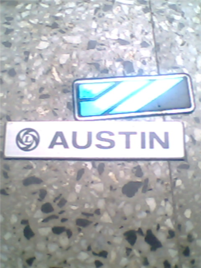 New Caterham Badge. - Page 1 - Caterham - PistonHeads - The image shows a top-down view of two license plates placed on a rough tiled surface. The larger plate, displaying the word "AUSTIN," has a black border and is oriented horizontally. The smaller plate, tightly packed and featuring a blue rectangle with white horizontal lines inside, is positioned vertically to the left of "AUSTIN." Both plates appear to be a part of a single vehicle, and the plates are of different shapes and colors. The overall impression is that of a license plate comparison.