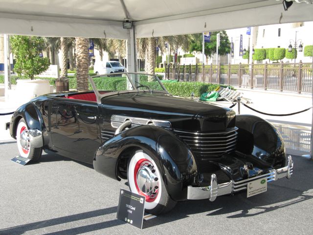 Emaar Pistonheads Classic - The image presents an antique car, likely a model from the early to mid-20th century, exhibited at an outdoor auto show or similar event. The car is predominantly black with some white detailing, and it features chrome elements that add a touch of classiness. It's equipped with whitewalls tires, a characteristic wheel design from earlier times. The setting is adorned with palm trees, and the car is stationed under a tent roof, suggesting a sunny day at the exhibit. The overall atmosphere of the scene is elegant and nostalgic, showcasing a piece of automotive history.