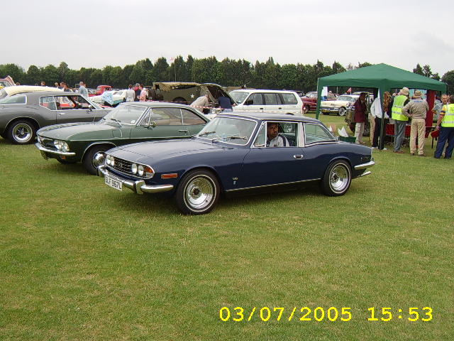 The image depicts a classic car show on a grassy field. There are two classic cars prominently displayed, a dark blue car with a hardtop convertible top and a lighter green classic car with a soft top. Several people can be seen, likely attendees and enthusiasts, admiring the classic cars. There's also a tent in the background, possibly providing shelter or serving as an information or merchandise booth. The overall atmosphere of the image is lively, with the focus on the classic vehicles and the people enjoying the event.