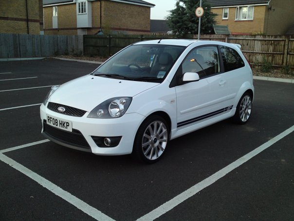 *STOLEN* Swansea/Caswell Bay/South Wales - Page 1 - South Wales - PistonHeads - The image showcases a white four-door hatchback compact car parked in a parking lot. The car is clean and well-maintained, with a visible licence plate that reads "B50 HKP." A secondary plate, presumably a registration marks, is mounted on the back. The vehicle's body exhibits a glossy finish, reflecting the surroundings, indicating that it is parked in a designated space, which is bordered by white lines and a white curb. In the background, there are residential buildings with a green lawn, suggesting that this lot might be near a residential area.