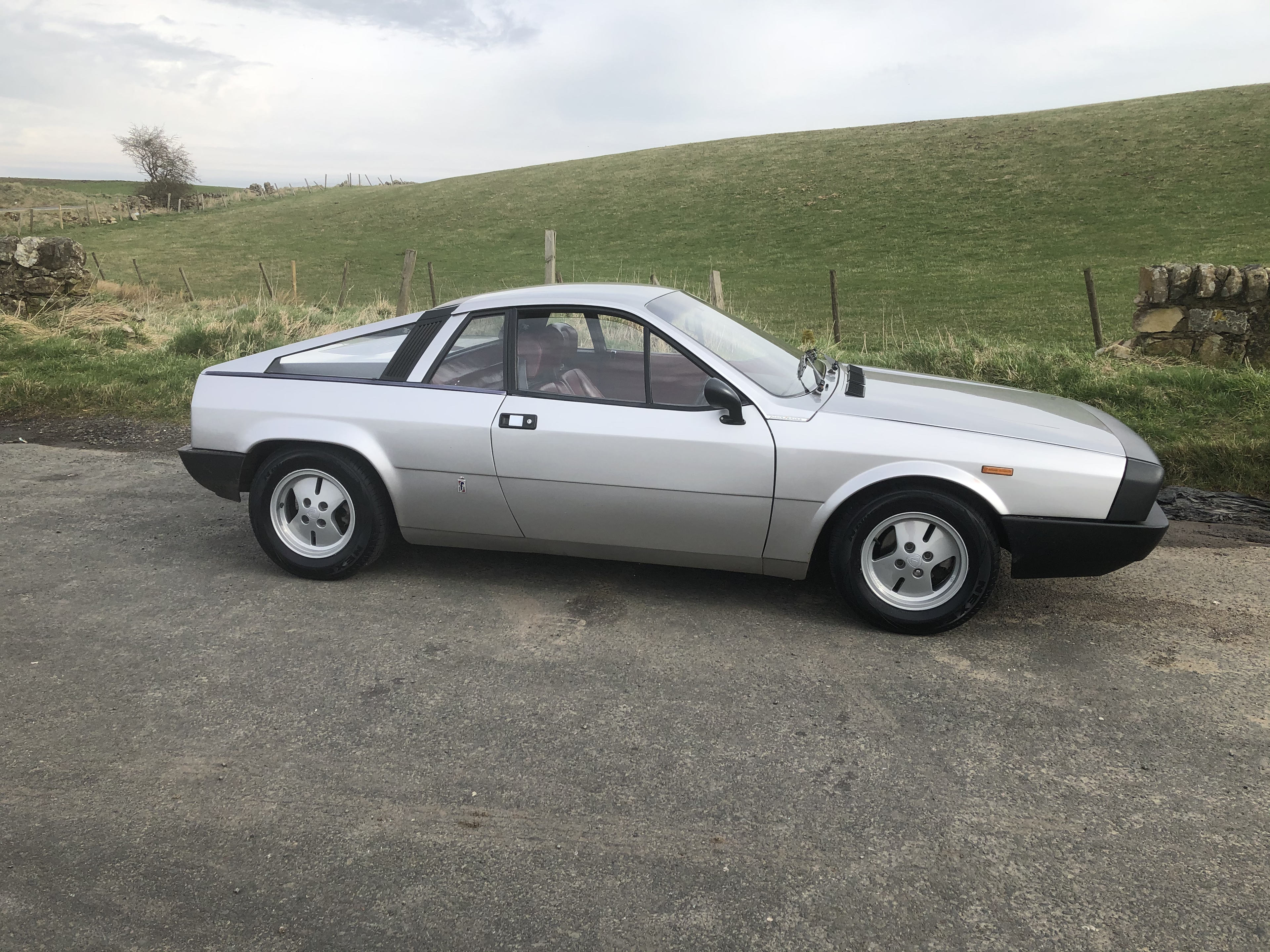 Lancia Montecarlo...37 years and counting - Page 13 - Readers' Cars - PistonHeads UK - The image features a vintage sports car parked on the side of a road. The car appears to be in good condition, with its distinctive white bodywork and unique design details that suggest it's a model from the 1980s or 1990s. It has a large rear wing and a prominent front spoiler, typical features of high-performance cars from that era. The car is positioned at an angle to the camera, showing its right side profile. There are no people visible in the immediate vicinity of the car, but there's a person visible in the background, suggesting activity beyond the car itself. The setting seems to be a rural or semi-rural area, as indicated by the open field and the stone wall in the distance.