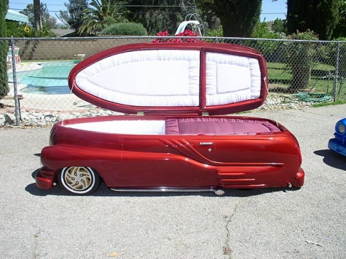 Would you be seen dead in this? - Page 1 - General Gassing - PistonHeads - The image presents an intriguing scene featuring a vintage car trailer that has been creatively repurposed into a coffin-shaped seating. The trailer, with its vibrant red and white hue, is parked in front of a chain-link fence, providing a stark contrast to its surroundings. The bed of the trailer is uniquely shaped like a coffin, complete with a white interior that stands out against the red exterior. Interestingly, the lid of the coffin is open, revealing an interior that mirrors the lid's design. The trailer is the sole object in the image, drawing the viewer's attention immediately to it. The overall composition of the image suggests a blend of macabre humor and creative reimagining of everyday objects.