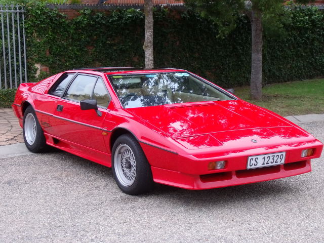 Greetings from a South African Esprit lover. - Page 1 - Esprit - PistonHeads - The image features a vibrant, glossy red car sitting on a gravel driveway. The car is a two-door sports model, possibly an Abarth, judging by its short and wide body. It has black trim around the headlights, which are visible on the front. The vehicle is positioned at an angle that shows its front left and rear right sides. The background is mostly made up of well-manicured greenery and a fence, creating a pleasant, suburban setting. The overall impressions suggest a personal or private ownership by someone with a distinctive taste for fast, stylish cars.