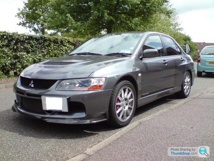 Post your Evo pics - Page 4 - Jap Chat - PistonHeads - The image showcases a dark-colored Mazda RX-8 sports car parked on a two-tone sidewalk. The car is positioned at an angle, facing slightly towards the right side of the image. Its wheels are silver alloy, matching its black trim. In the background, there's a glimpse of a green car and lush greenery, adding a touch of nature. The photograph seems to be taken during the day under bright sunlight, as evidenced by shadows cast on the ground.