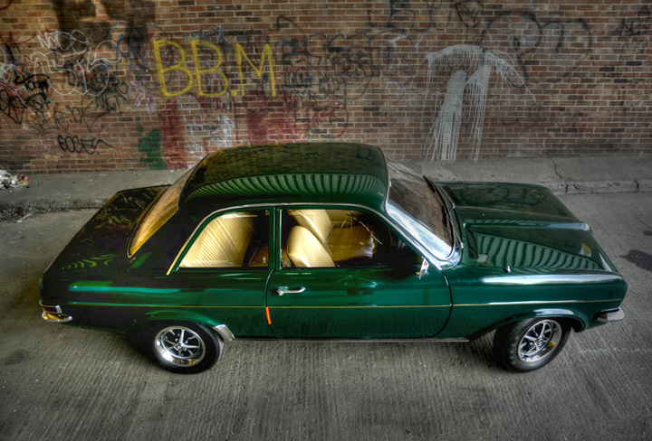 Pistonheads Classics - The image features a dark green classic car parked on a concrete surface. The vehicle displays a shiny exterior, likely due to its use of a 2K cleaner, as indicated on the side of the car. The background shows a brick wall with various graffiti, including letters and abstract designs, which contribute to an urban or street art atmosphere.